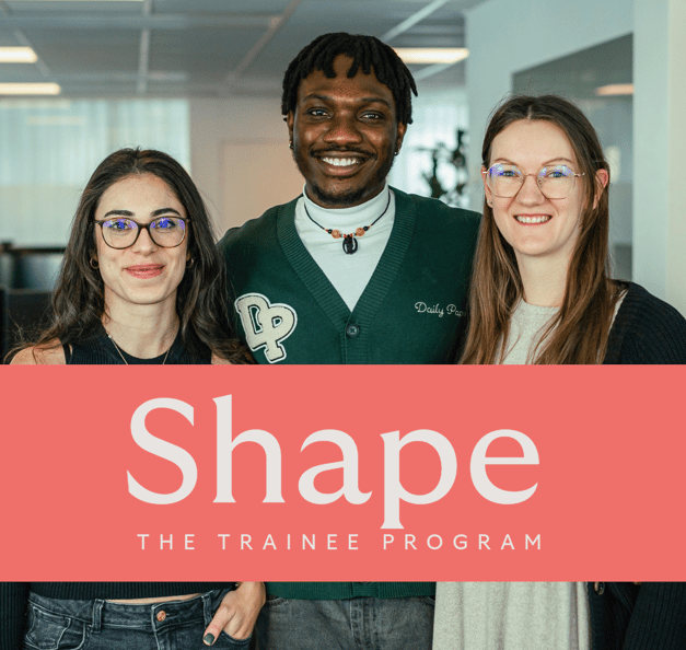 Three smiling young colleagues standing side by side in a bright office. A coral banner across the image reads "Shape – The Trainee Program".