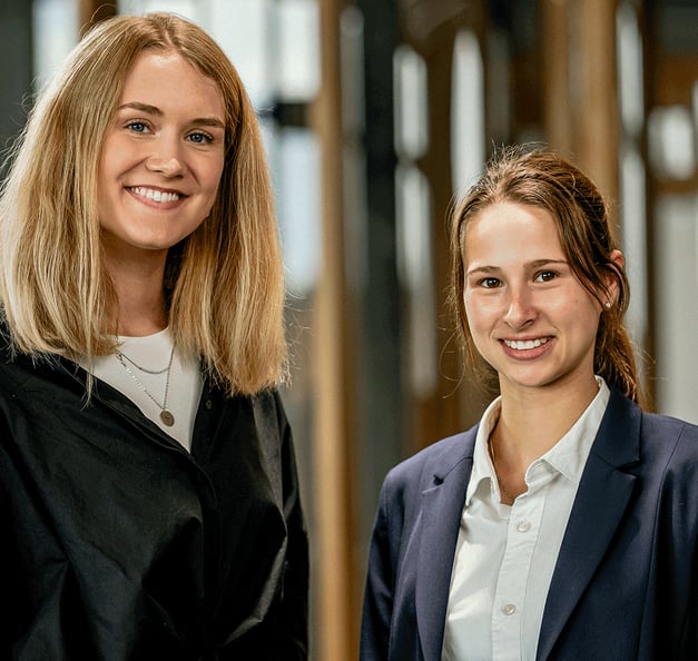 Two smiling young colleagues standing side by side in a modern office with wooden frames and glass walls – one wearing a black blouse, the other a navy blazer over a white shirt.