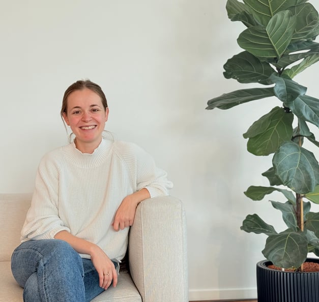 A woman sitting on a beige sofa, smiling warmly at the camera. She is wearing a white sweater and blue jeans, with a large green potted plant placed beside her against a plain white wall.
