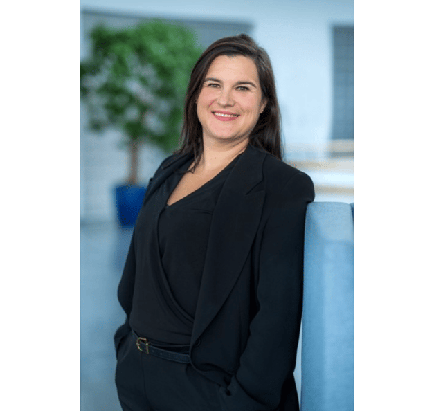 A woman with dark hair is smiling and standing indoors, wearing a black outfit and blazer. The background is softly blurred, showing a modern office space with a plant in the distance.