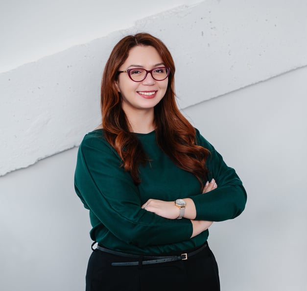 A woman with long brown hair and glasses is smiling with her arms crossed, wearing a dark green top and standing against a light-colored background.