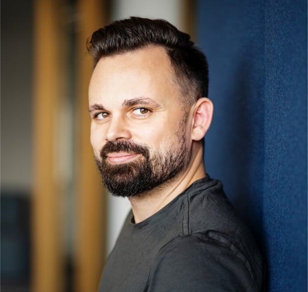 A man with short dark hair and a beard is standing indoors, leaning slightly against a blue wall and looking at the camera with a slight smile. The background is softly blurred.
