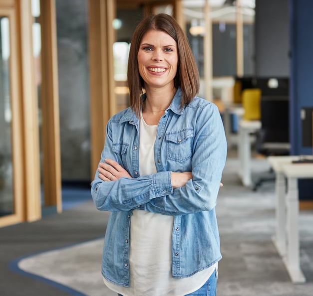 Woman with shoulder-length brown hair wearing a denim shirt and white top, standing with arms crossed and smiling in a modern office environment.