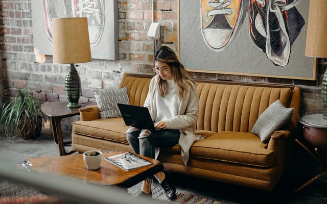 Eine junge Frau sitzt auf einem senffarbenen Sofa und schaut auf ihren Laptop