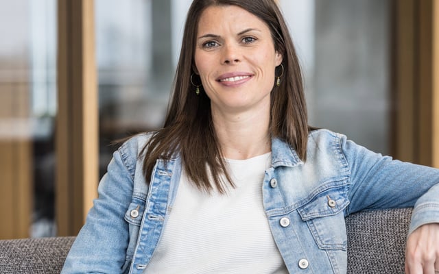 The image shows Carina seated in a relaxed posture on a grey upholstered sofa or armchair. The person has long, straight, dark brown hair worn open, falling onto their shoulders. Their facial expression is friendly and open: they are smiling naturally into the camera, conveying approachability and ease. The person is wearing a white top with a fine texture and a light blue denim jacket over it. Small green earrings are visible as accessories. One arm rests loosely on the back of the seat, emphasising the relaxed body posture. The background shows a modern, bright office or workspace with large windows through which natural daylight enters. The environment appears professional and welcoming. No text, logos, technologies, or accessibility symbols are visible in the image. The photograph is composed as a professional portrait, likely intended for a professional or institutional context.