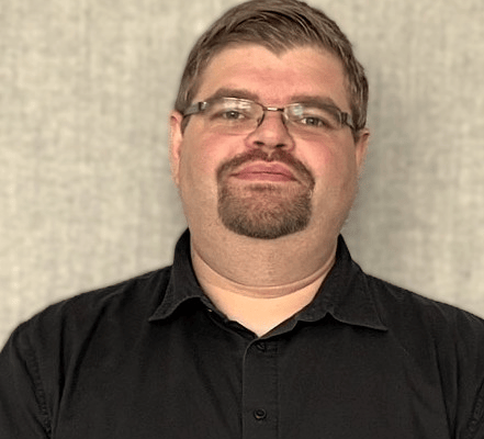 A person is standing in front of a neutral, textured grey background. They are wearing a short-sleeved black button-up shirt and a dark smartwatch on their left wrist. Their arms are loosely folded in front of their body. The person has short brown hair, wears thin-framed metal glasses, and has a neatly trimmed goatee. Their facial expression appears calm and friendly. No branding or accessibility-relevant features are visible in the image.
