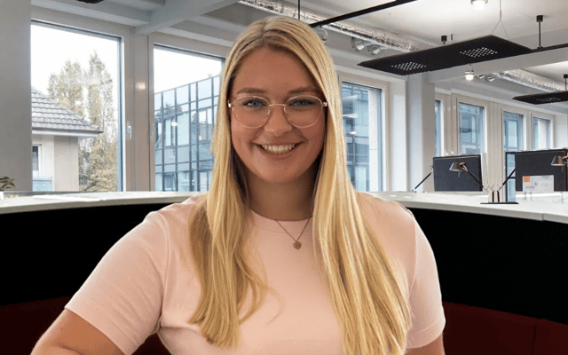 Portrait of a young professional woman with long blonde hair, smiling in an office environment, wearing a black v-neck top and a necklace with a letter pendant.