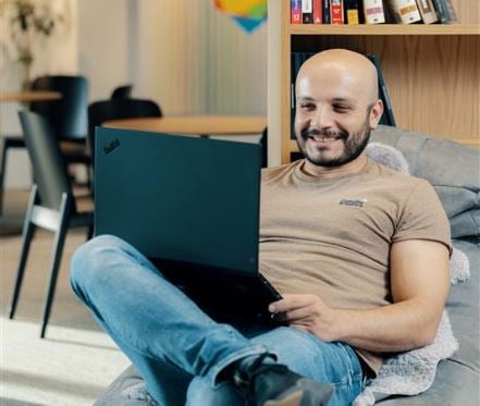 Man sitting comfortably on a beanbag, smiling while using a laptop in a modern office setting