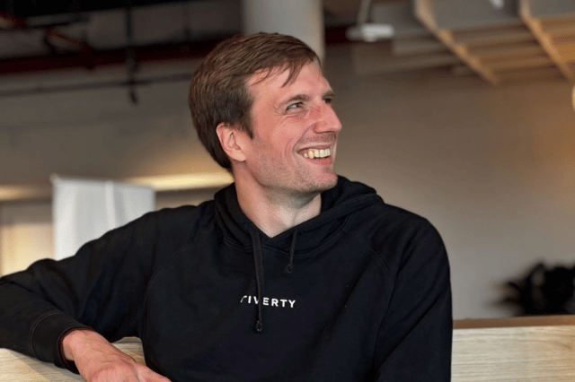 An employee in a black hoodie with the Riverty logo laughs and looks to the side, holding a coffee mug, with a bright open-plan office in the background.