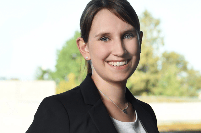 Portrait of a smiling woman with dark hair tied back, wearing a black blazer over a light top, standing outdoors in front of blurred trees.