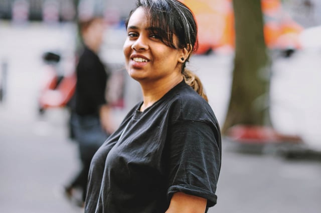 Smiling woman standing outdoors on a city street, wearing a dark t-shirt, with blurred people and trees in the background