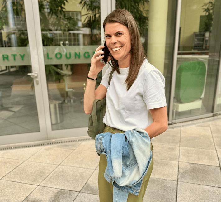 The image shows a person standing outdoors in front of a modern building while talking on the phone. The person has long, straight, dark brown hair worn open. Their facial expression is very cheerful and open: they are laughing broadly into the camera, conveying energy and a positive mood. The person is wearing a plain white t-shirt and olive green trousers. A light blue denim jacket is held loosely in one hand rather than being worn. An olive green shoulder bag is visible over one shoulder. A black bracelet is visible on the wrist. In one hand, the person is holding a dark smartphone pressed to their ear. In the background, a glazed building facade is visible with green lettering, of which the words "RIVERTY" and "LOUNGE" are legible. Through the glass panels, plants and seating furniture are visible inside the building. The ground consists of light stone tiles. The surroundings appear urban and publicly accessible.