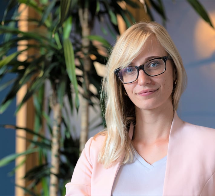 A blonde woman with black-rimmed glasses wearing a light pink blazer and grey top, looking at the camera with a subtle smile, set against an indoor background with leafy green plants.