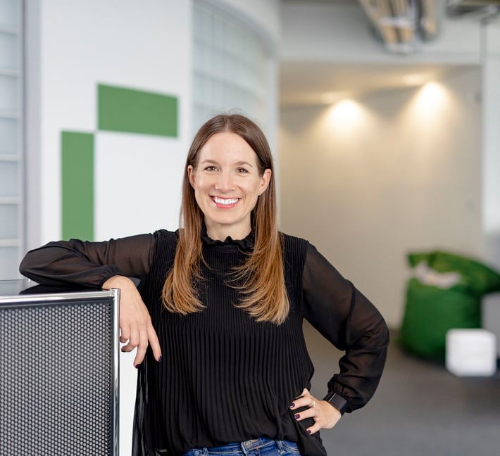 A woman with long brown hair is smiling and standing in a modern office space, leaning casually on a metal cabinet. She is wearing a black blouse and jeans, with soft lighting and green accents in the background.