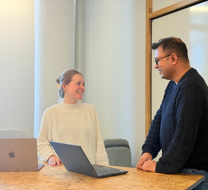 Zwei Personen stehen an einem Holztisch in einem hellen Besprechungsraum, lächeln und unterhalten sich miteinander. Vor ihnen steht jeweils ein offener Laptop, und an der Wand hinter ihnen ist ein Whiteboard zu sehen.