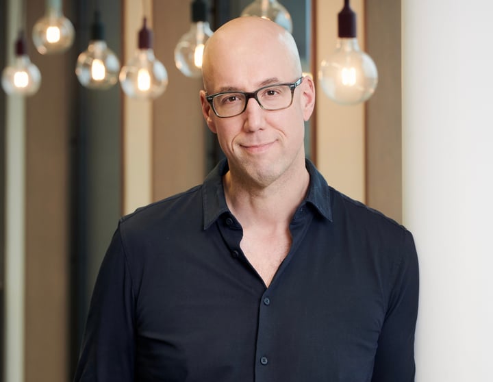 Professional portrait of a man in casual business attire standing in a modern office environment with soft lighting and a blurred background.