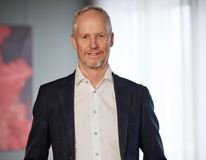 Porträtt av en man i mörk kavaj och vit skjorta, stående inomhus.