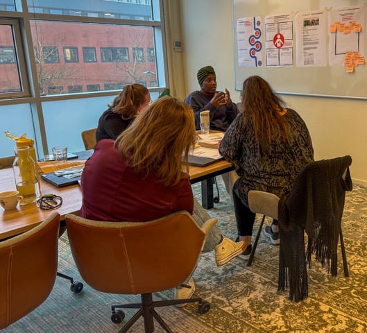 Workshopsessie bij Riverty waarbij deelnemers rond een tafel zitten en luisteren naar een ervaringsdeskundige van Stichting ONSbank, met notities en posters over jongeren en financiën zichtbaar aan de muur in een vergaderruimte.