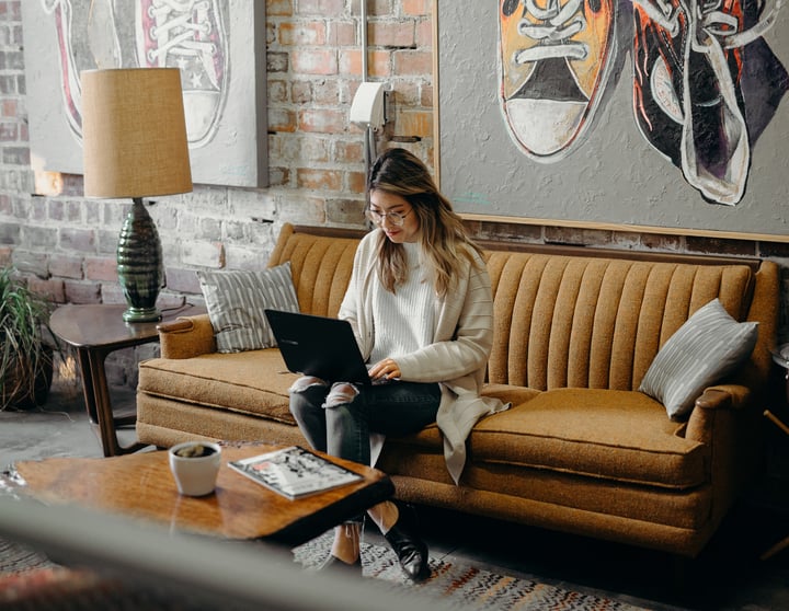 Eine junge Frau sitzt auf einem senffarbenen Sofa und schaut auf ihren Laptop