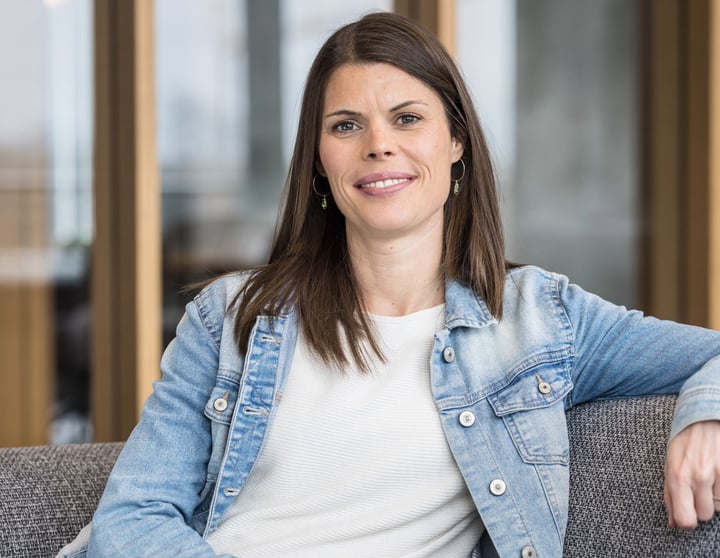 The image shows Carina seated in a relaxed posture on a grey upholstered sofa or armchair. The person has long, straight, dark brown hair worn open, falling onto their shoulders. Their facial expression is friendly and open: they are smiling naturally into the camera, conveying approachability and ease. The person is wearing a white top with a fine texture and a light blue denim jacket over it. Small green earrings are visible as accessories. One arm rests loosely on the back of the seat, emphasising the relaxed body posture. The background shows a modern, bright office or workspace with large windows through which natural daylight enters. The environment appears professional and welcoming. No text, logos, technologies, or accessibility symbols are visible in the image. The photograph is composed as a professional portrait, likely intended for a professional or institutional context.