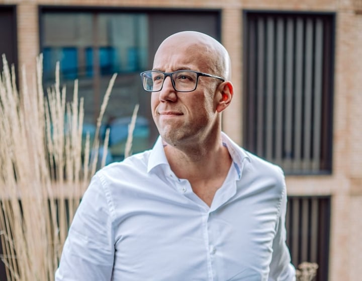 Kale man met een bril en een wit overhemd die buiten staat en bedachtzaam opzij kijkt, met een modern gebouw op de achtergrond