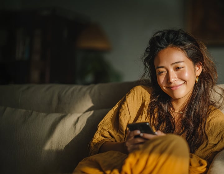 A young woman with long dark hair, dressed in a loose mustard-yellow outfit, sits comfortably on a couch, smiling as she looks at her smartphone. Warm sunlight softly illuminates her face, creating a cozy and relaxed atmosphere