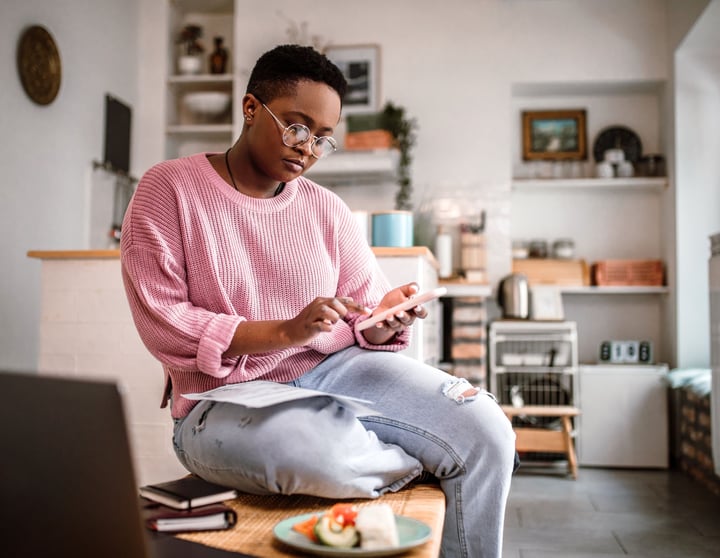 Eine junge Frau in einem rosa Pullover sitzt lässig auf ihrem Schreibtisch und schaut auf ihr Handy