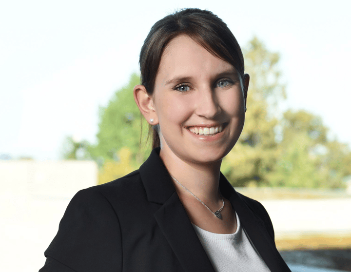 At the center of the image, a person is facing the camera and smiling warmly. The person is wearing a black blazer over a light-colored top. Their hair is brown, pulled back, with a slightly off-center part. Visible accessories include small stud earrings and a delicate necklace with a small leaf- or teardrop-shaped pendant. The background is blurred and shows trees and greenery outdoors, with a light horizontal area in the lower part of the image. No text, logos, or brand markings are visible. The overall impression is that of a professional portrait taken outside.