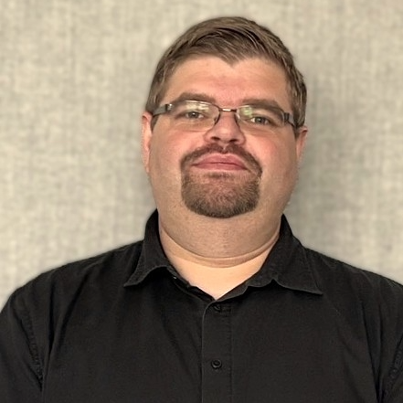 A person is standing in front of a neutral, textured grey background. They are wearing a short-sleeved black button-up shirt and a dark smartwatch on their left wrist. Their arms are loosely folded in front of their body. The person has short brown hair, wears thin-framed metal glasses, and has a neatly trimmed goatee. Their facial expression appears calm and friendly. No branding or accessibility-relevant features are visible in the image.