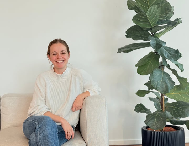 Eine lächelnde Person sitzt auf einem hellen Sofa, trägt einen weißen Pullover und Jeans, und neben ihr steht eine große, grüne Pflanze in einem Topf vor einer schlichten Wand.