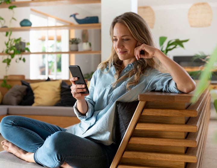 Eine Dame sitzt lächelnd auf der Couch und schaut auf ihr Telefon
