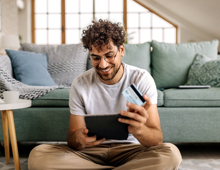 Ein junger Mann sitzt auf einem Teppich in seinem Wohnzimmer, schaut auf sein Tablet und hält eine Kreditkarte in einer Hand