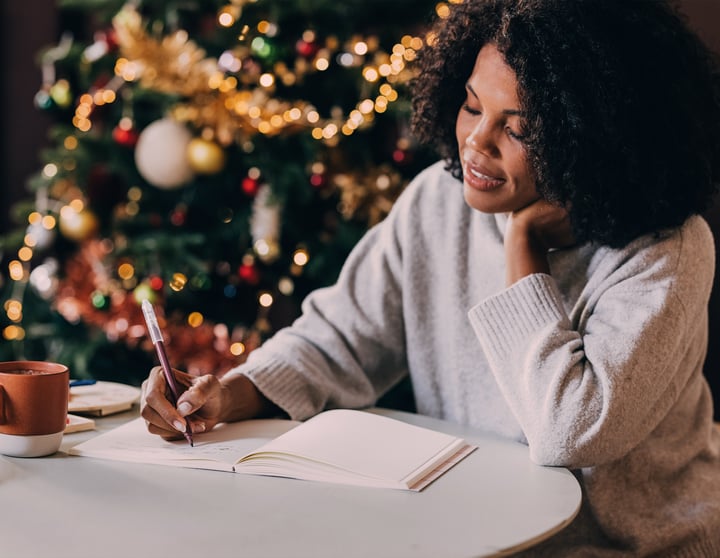 Eine Frau sitzt entspannt an einem Tisch vor einem festlich geschmückten Weihnachtsbaum, schreibt in ein Notizbuch und trinkt aus einer Tasse.