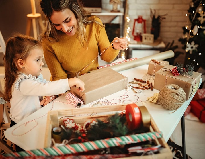Eine Mutter und ihre Tochter verpacken gemeinsam Weihnachtsgeschenke während im Hintergrund ein geschmückter Weihnachtsbaum für festliche Stimmung sorgt.