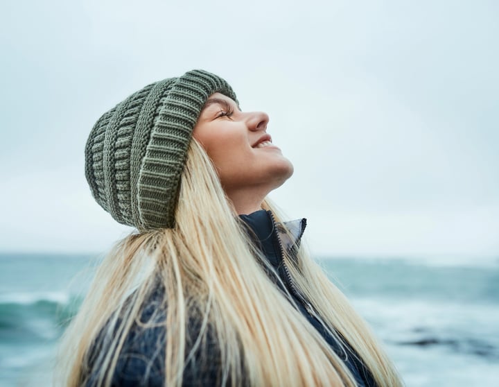 Eine junge blonde Frau hat ihren Kopf in den Nacken gelegt und ihre Augen verschlossen und genießt die kalte Brise am Meer