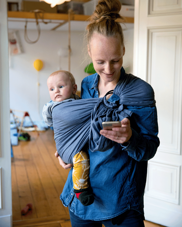 Junge Mutter, welche ihr Baby im Arm trägt und gleichzeitig auf ihr Smartphone schaut
